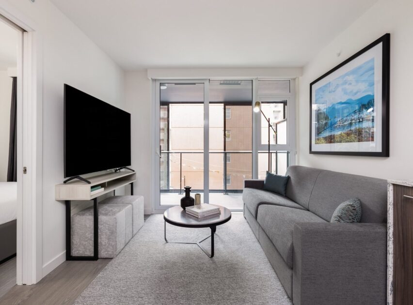 Modern living room with a gray sofa, round coffee table, large flat-screen TV on a console, light rug, framed mountain artwork, and floor-to-ceiling windows leading to a balcony with urban views.