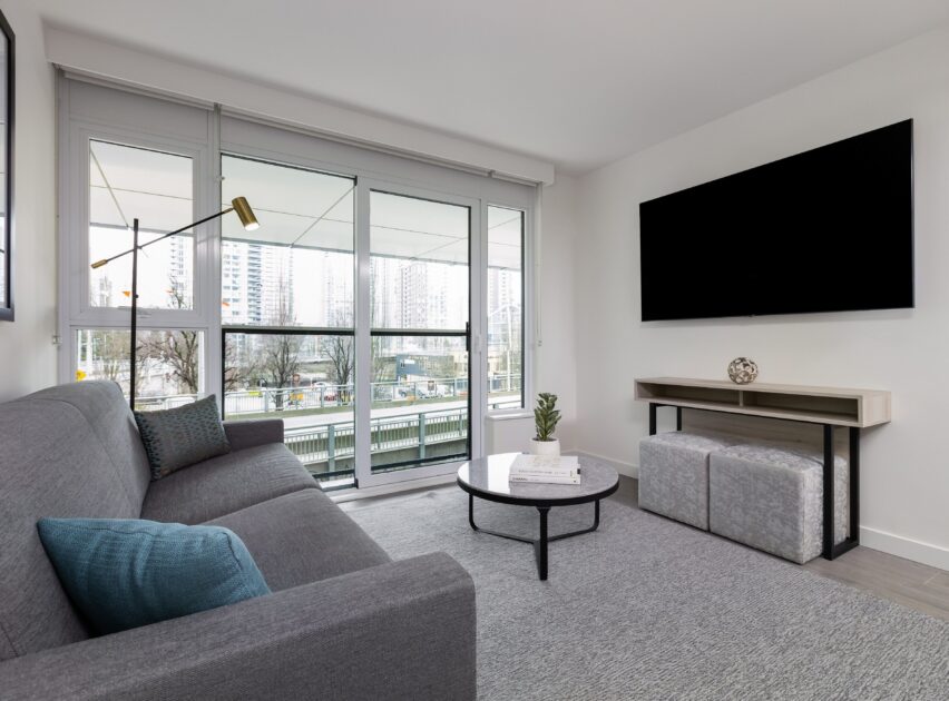 Modern living room with a gray sofa, blue accent pillow, round coffee table, and wall-mounted TV. Large windows provide city views. A console table with decor and two ottomans sits beneath the TV.