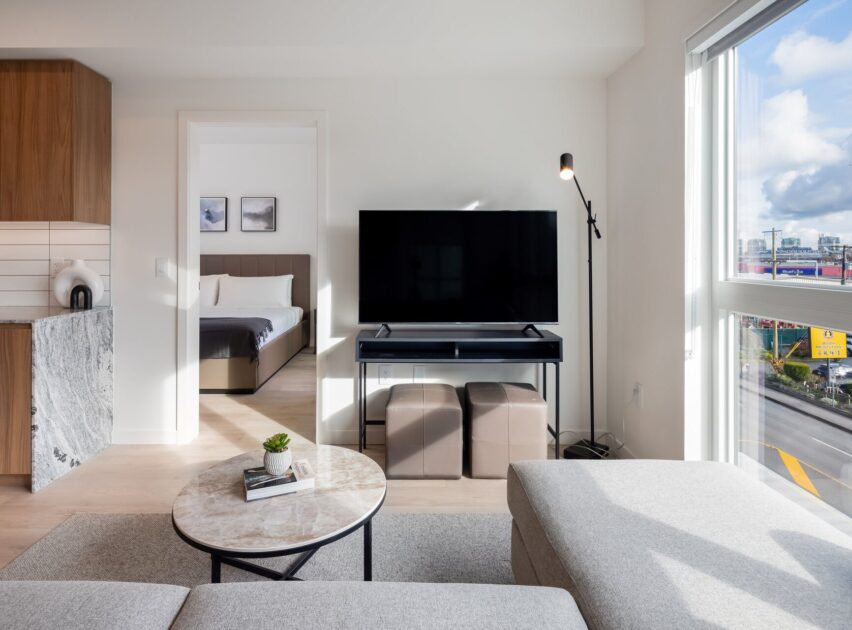 A modern living room with a grey sectional sofa, round coffee table, TV on a stand, and floor lamp. Large window shows a city view. An adjacent bedroom with a bed is visible through an open doorway.