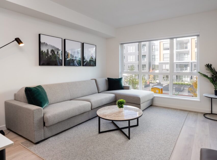 Modern living room with a light gray sectional sofa, green accent pillows, round marble coffee table, wall art, floor lamp, large window with natural light, and view of apartment buildings outside.