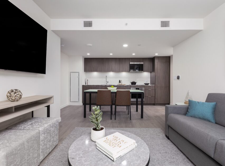 Modern open-plan living room and kitchen with gray sofa, blue cushion, round coffee table, two bar stools, dark wood cabinets, and neutral decor. A large wall-mounted TV is visible on the left wall.