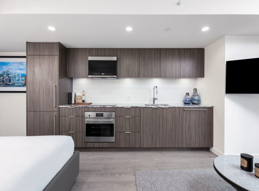 Modern studio kitchen with sleek wood cabinets, built-in oven, microwave, sink, and countertop decor. The space is well-lit with recessed ceiling lights and features a bed, wall art, and a mounted TV.