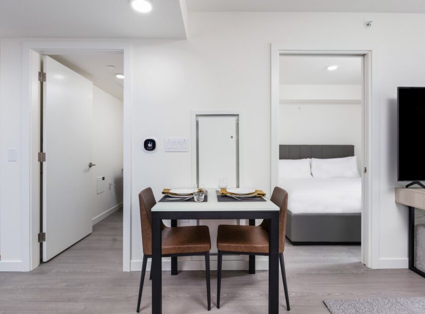 A modern apartment interior with a small dining table set for two, brown chairs, and a view into a bedroom with a neatly made bed and white bedding. The space features light-colored walls and flooring.