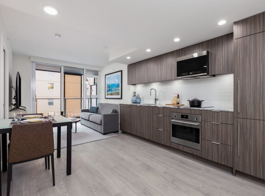 Modern open-plan kitchen and living area with wood cabinets, built-in appliances, dining table set for two, gray sofa, large window, and light wood flooring. Bright and tidy contemporary interior.