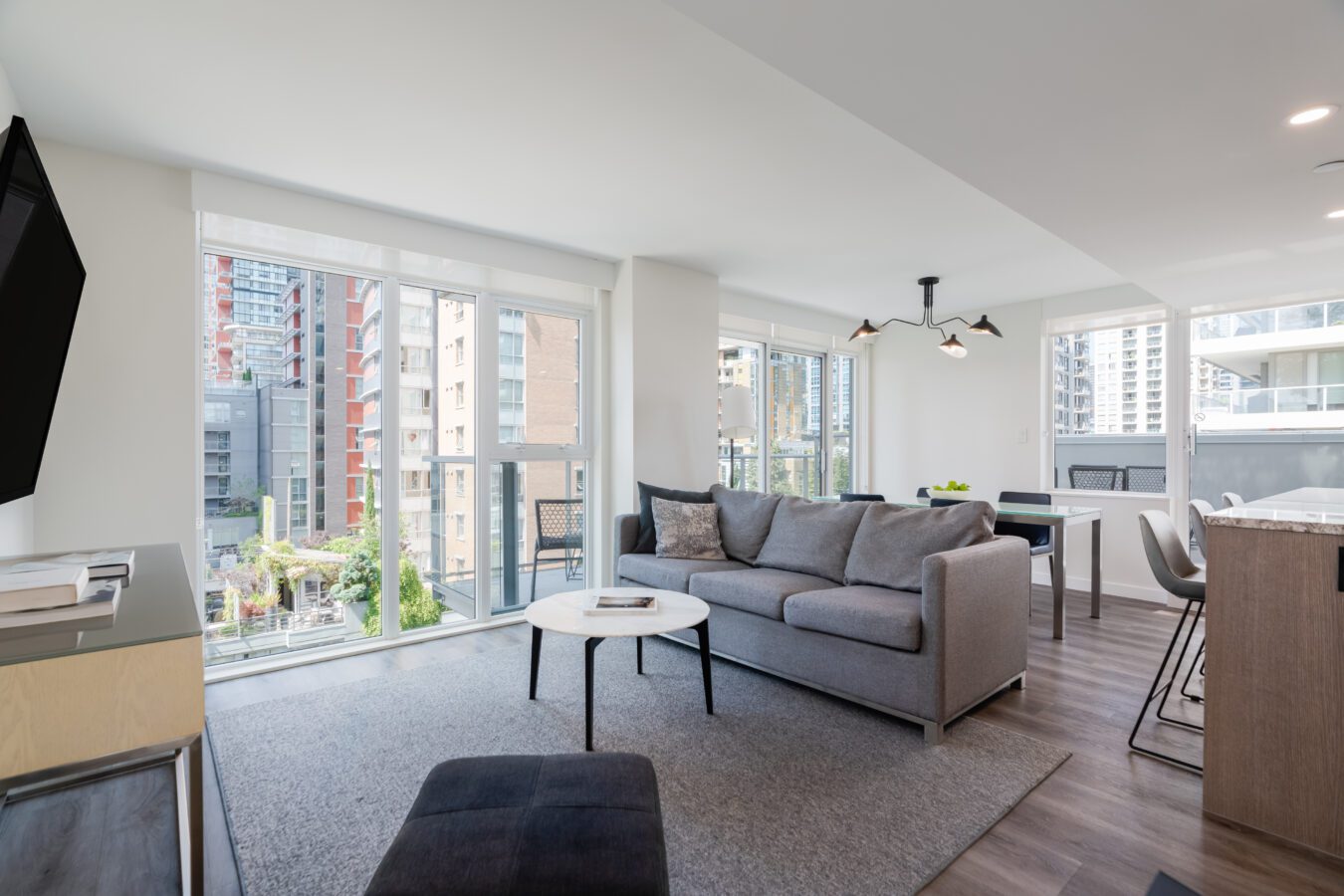 Modern, bright living room with large windows showing city views, a gray sofa, white round coffee table, gray rug, and an open kitchen and dining area with contemporary decor.