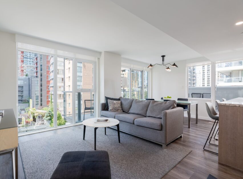 A modern, bright living room with large windows, a gray sofa, round white coffee table, dining area, and city views of tall buildings outside. The space has light walls and hardwood floors.