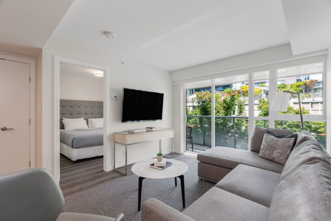 Modern living room with a gray sectional sofa, round coffee table, TV mounted on the wall, and large windows letting in natural light. A bedroom with a made-up bed is visible through an open doorway.