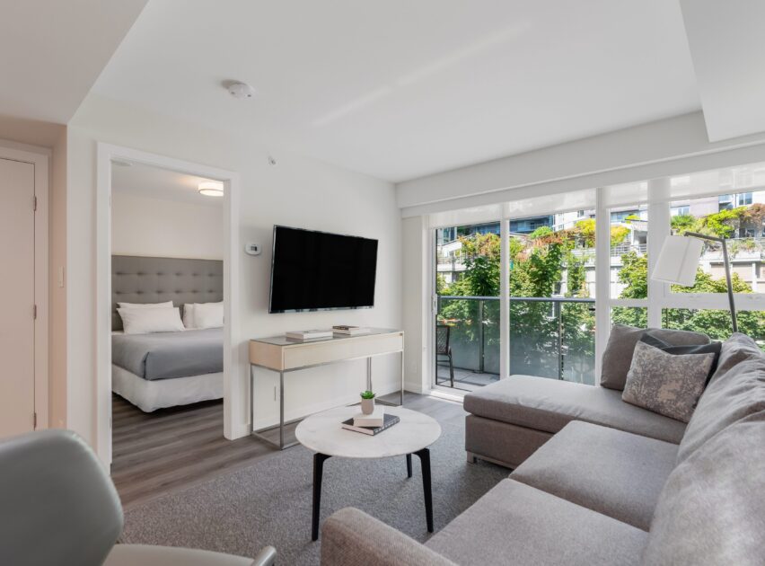 Modern living room with a gray sectional sofa, round coffee table, wall-mounted TV, and large window. Bedroom with a bed is visible through an open door. Natural light fills the space; greenery seen outside.