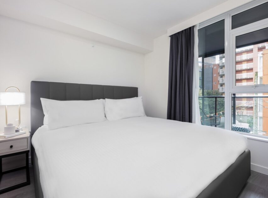 A modern, minimalist bedroom with a large bed, white bedding, a nightstand with a lamp and cups, and floor-to-ceiling windows with dark curtains, overlooking a cityscape with buildings.
