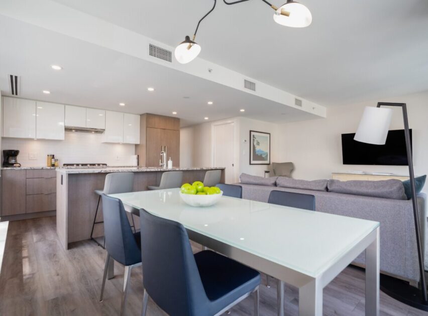 Modern open-concept kitchen and living room with a glass dining table, blue chairs, a bowl of green apples, gray sofas, flat-screen TV, and contemporary lighting, featuring neutral tones and wood flooring.