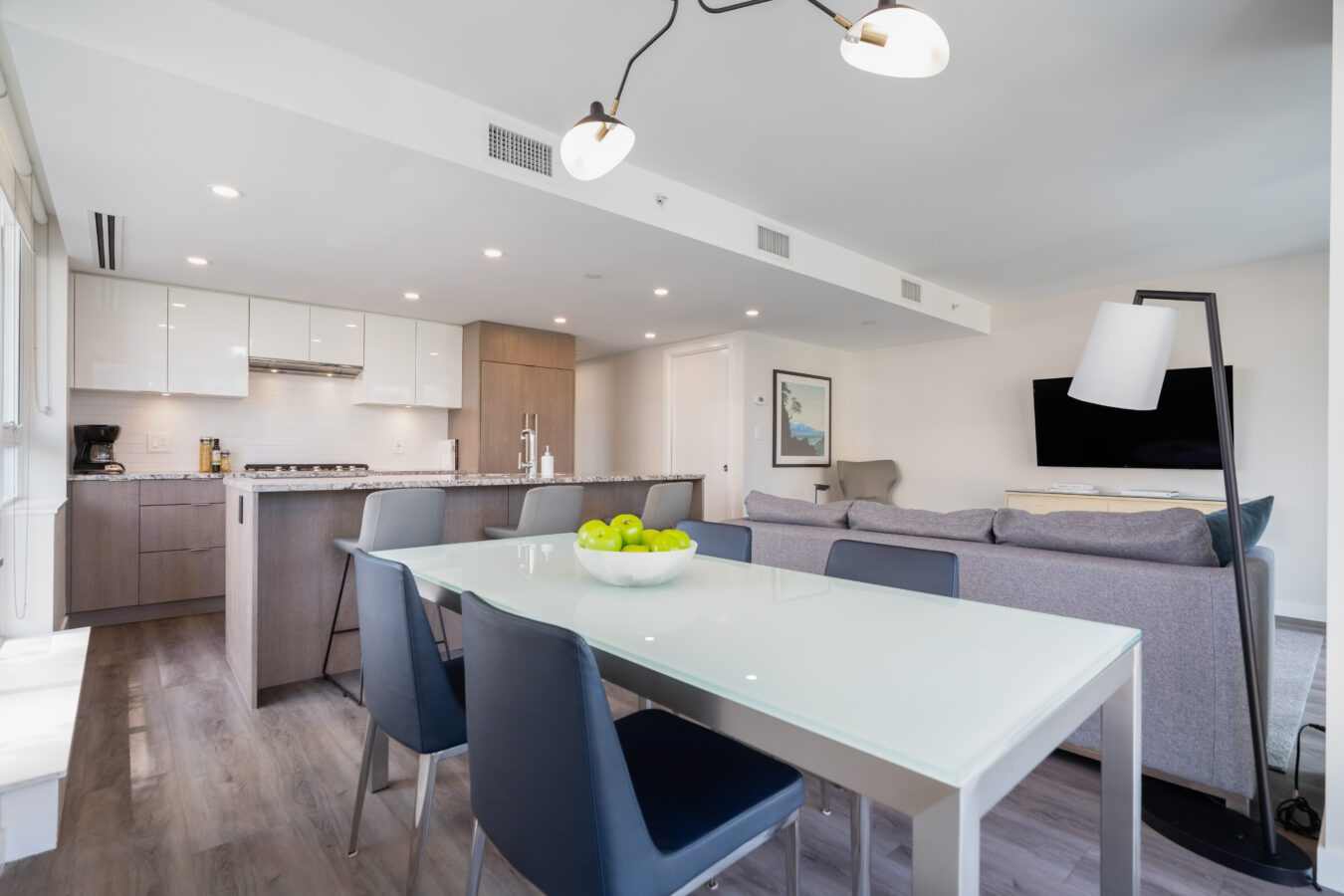 Modern open-concept kitchen and living room with a glass dining table, dark chairs, gray sofa, wall-mounted TV, and a bowl of green apples as a centerpiece. Bright, minimalist decor with wood floors.