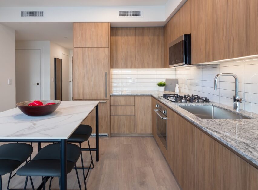 A modern kitchen with wood cabinets, a marble countertop island with black stools, stainless steel appliances, a built-in fridge, gas stove, and white subway tile backsplash. A bowl with red apples sits on the island.