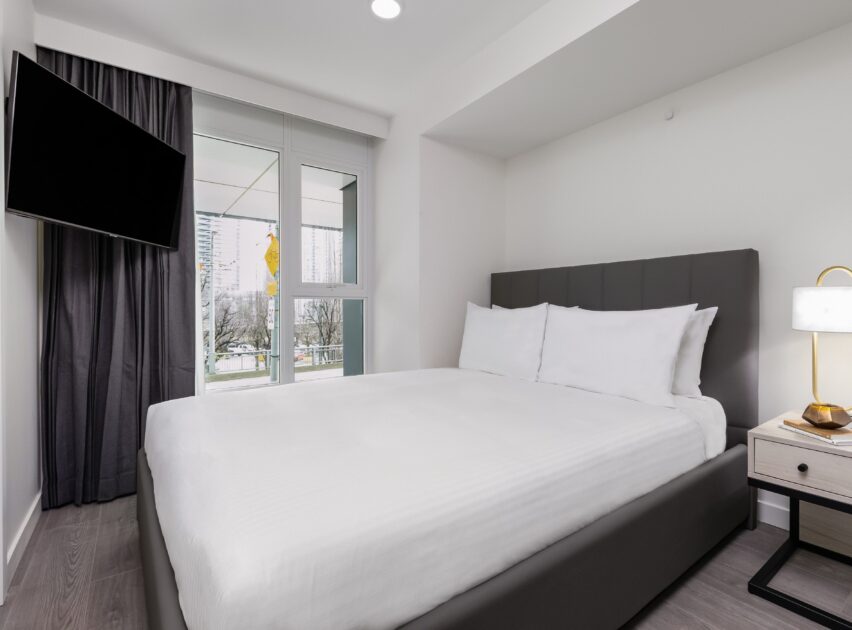 A modern, minimal bedroom with a large bed, white bedding, a bedside table and lamp, wall-mounted TV, dark curtains, and windows showing an urban view with trees and buildings.