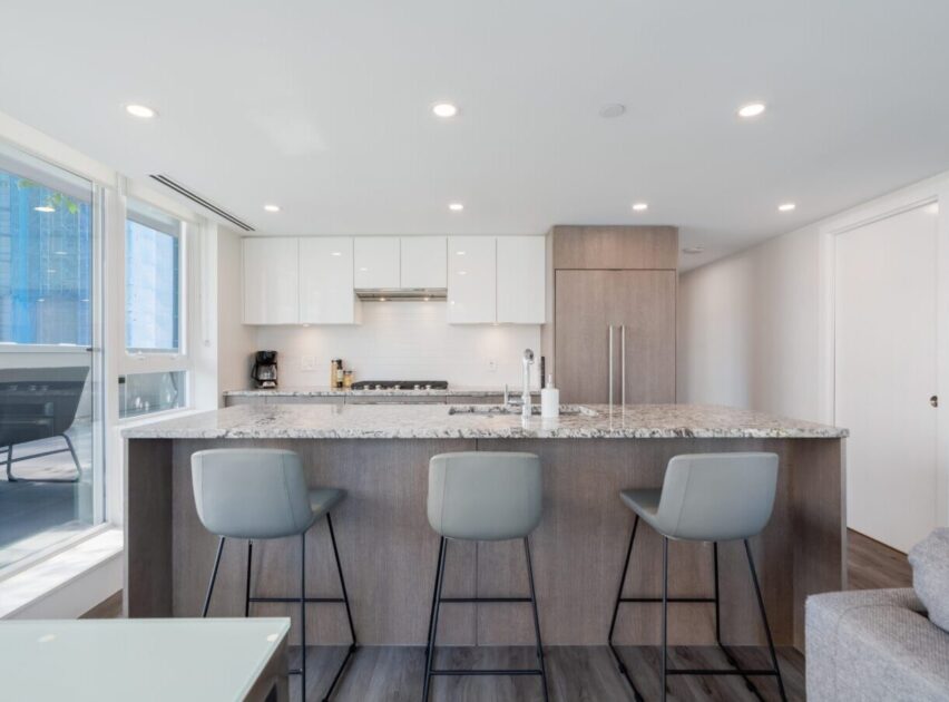 Modern kitchen with a large island featuring a granite countertop, three gray barstools, white cabinets, built-in appliances, and bright natural light from large windows.