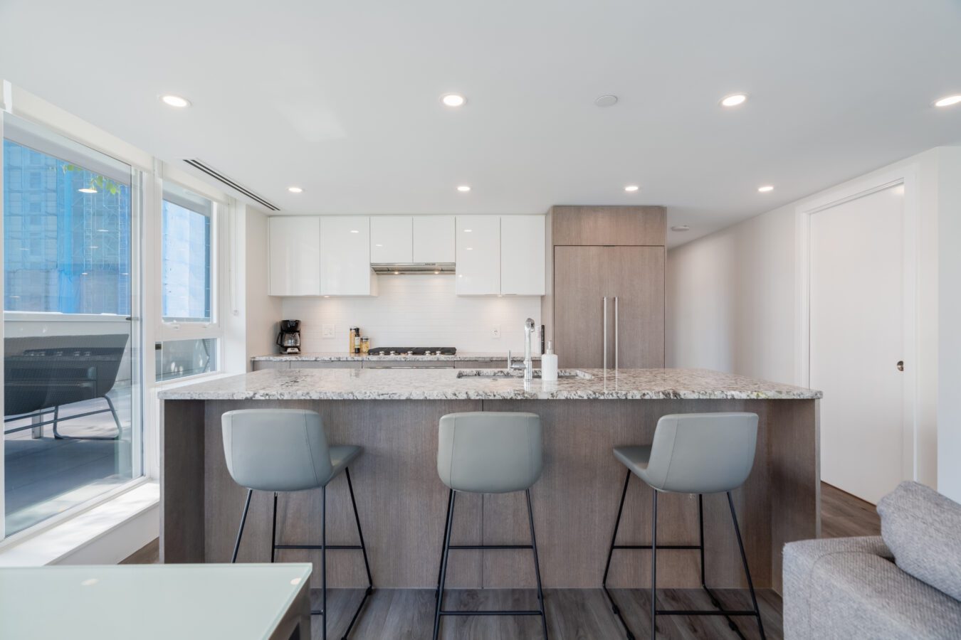 Modern kitchen with a large granite island, three gray barstools, white cabinets, built-in appliances, and bright lighting. Floor-to-ceiling windows provide natural light and a city view.