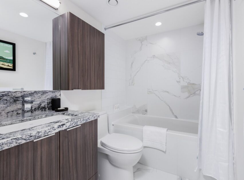 Modern bathroom with a wood-paneled vanity, marble countertop, white toilet, bathtub with marble tile wall, and a white shower curtain. A framed picture hangs on the white wall above the vanity.