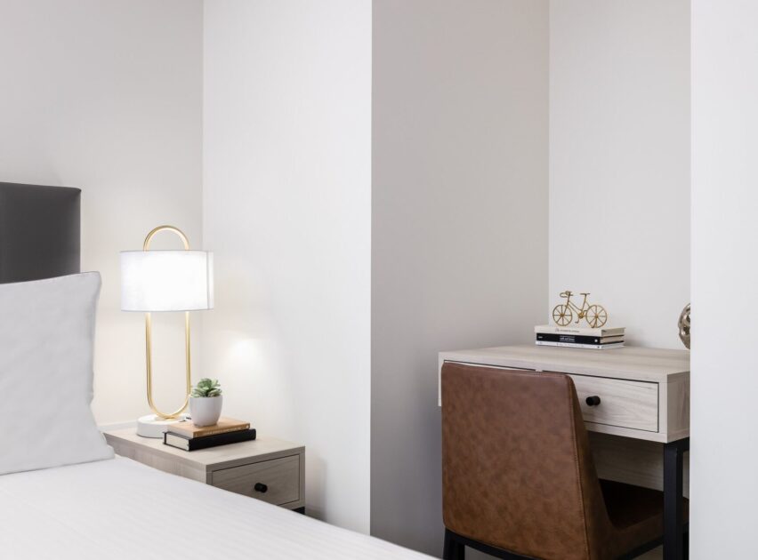 A modern, minimalist bedroom features a bed with white linens, a nightstand with a lamp and small plant, and a desk with a brown chair, stacked books, and a bicycle decor piece.