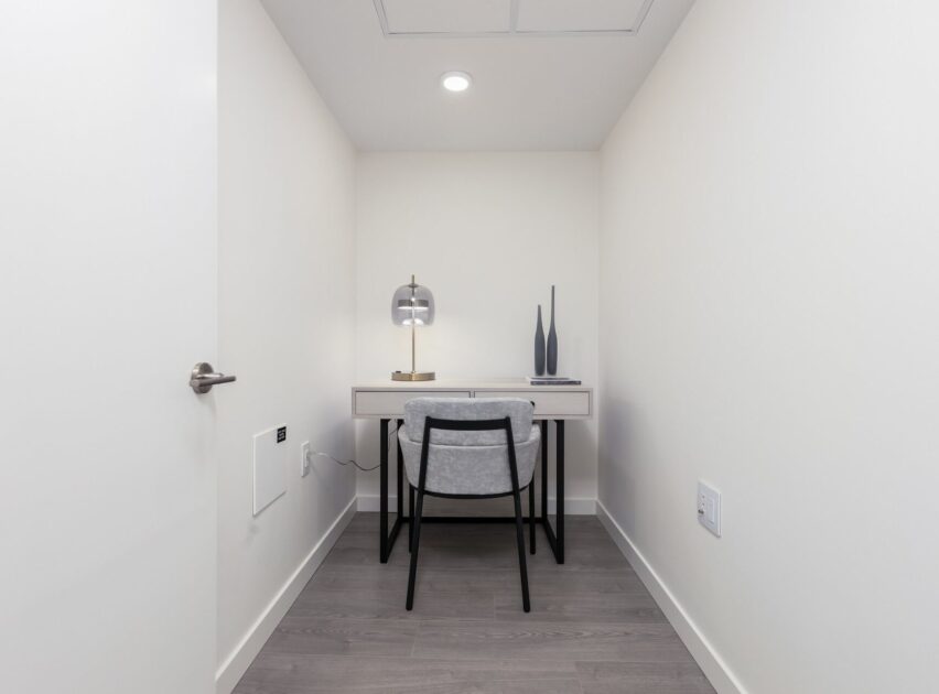 A minimalist home office with a small desk, a chair, a lamp, and two decorative vases against a plain white wall. The room has light-colored walls and gray wood flooring, creating a clean and simple workspace.