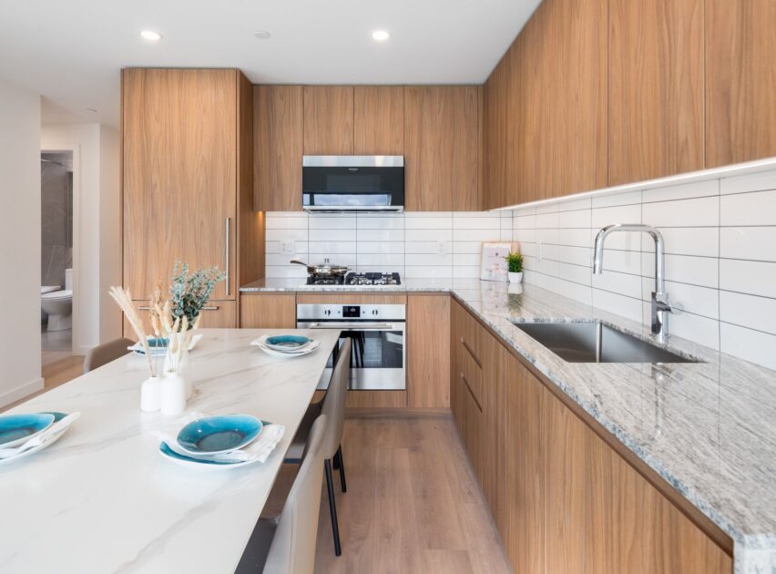 Modern kitchen with light wood cabinets, white countertops, stainless steel appliances, a gas stove, and a sink. A dining table set for four with blue plates is in the foreground. White tile backsplash on the wall.