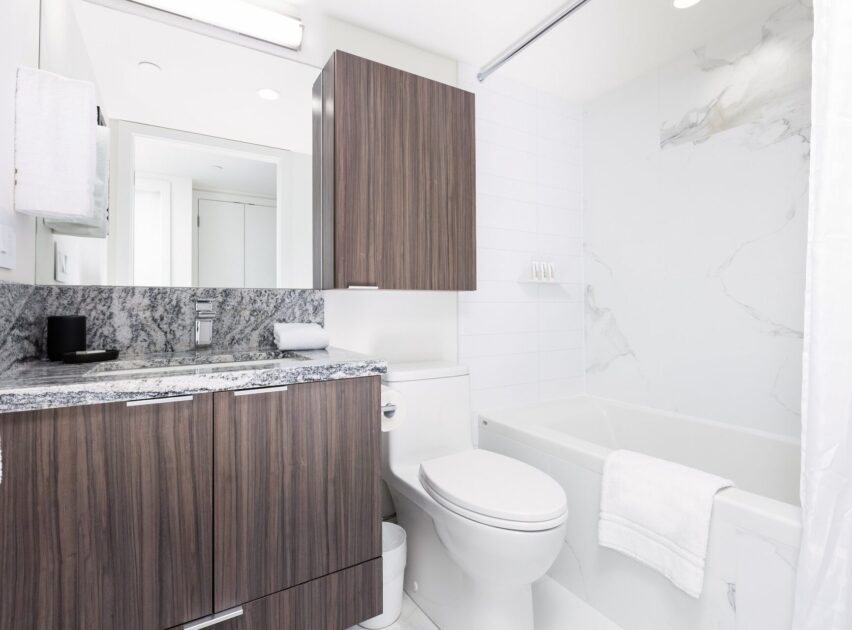 Modern bathroom with a wooden vanity, granite countertop, toilet, and a bathtub with white marble-style walls. A large mirror and bright lighting create a clean, minimalist look. White towel hangs on the bathtub.