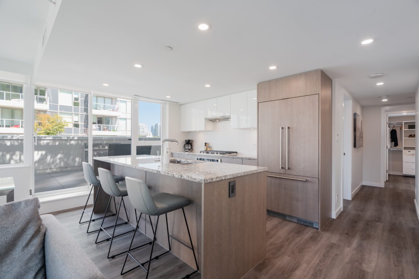 Modern kitchen with a large island, three gray barstools, light wood cabinets, stainless steel appliances, and floor-to-ceiling windows leading to a balcony with city views.