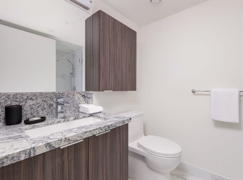 Modern bathroom with a granite countertop, wood cabinets, white sink, toilet, a wall mirror, and a towel hanging on a rack. Minimalist decor with folded white towels and a black soap dispenser.
