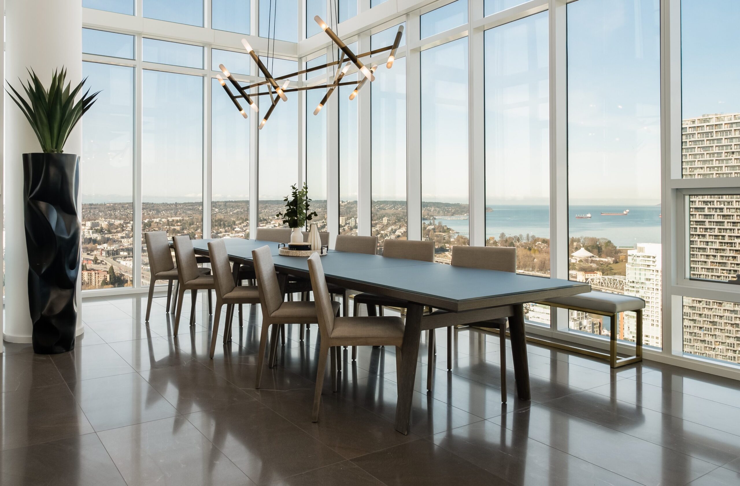 Modern dining room with a long table and ten chairs, large windows providing panoramic city and water views, a geometric chandelier, and a potted plant in the corner.