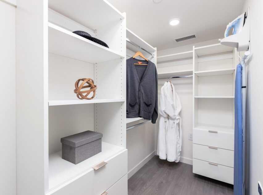 Walk-in closet with white shelves, drawers, and hanging rods. A gray sweater, a white robe, a hat, a decorative object, a gray storage box, and an iron with ironing board are visible in the organized space.