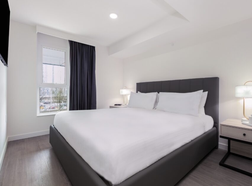 A modern, minimalistic bedroom with a large bed featuring white linens, two bedside tables with lamps, a window with dark curtains, and light-colored walls and flooring.
