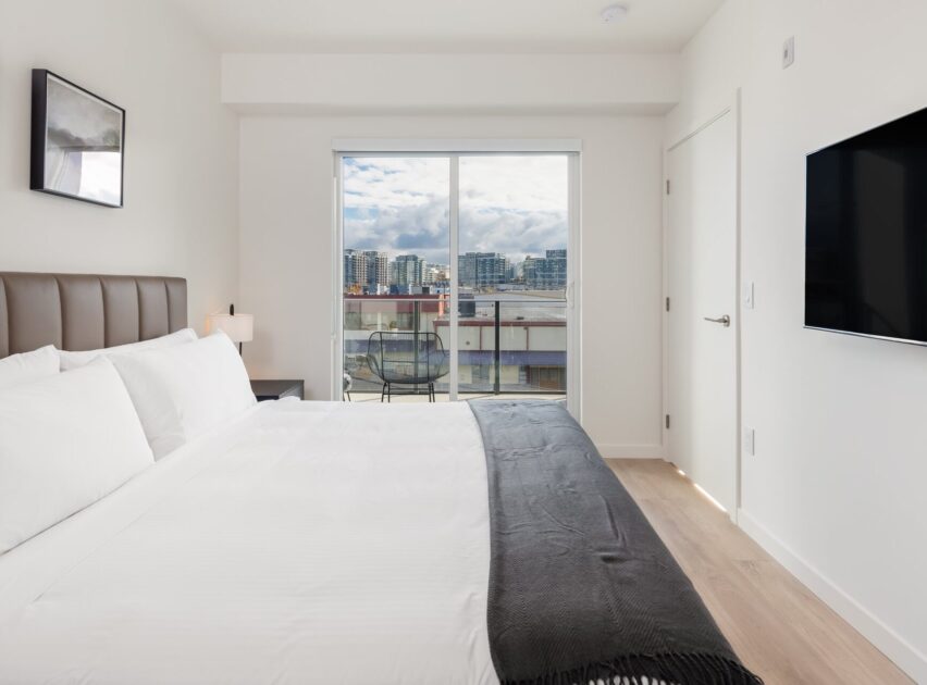 A modern, minimal bedroom with a large bed, white bedding, and a dark throw blanket. There’s a wall-mounted TV, a small nightstand with a lamp, and glass doors leading to a balcony overlooking city buildings.