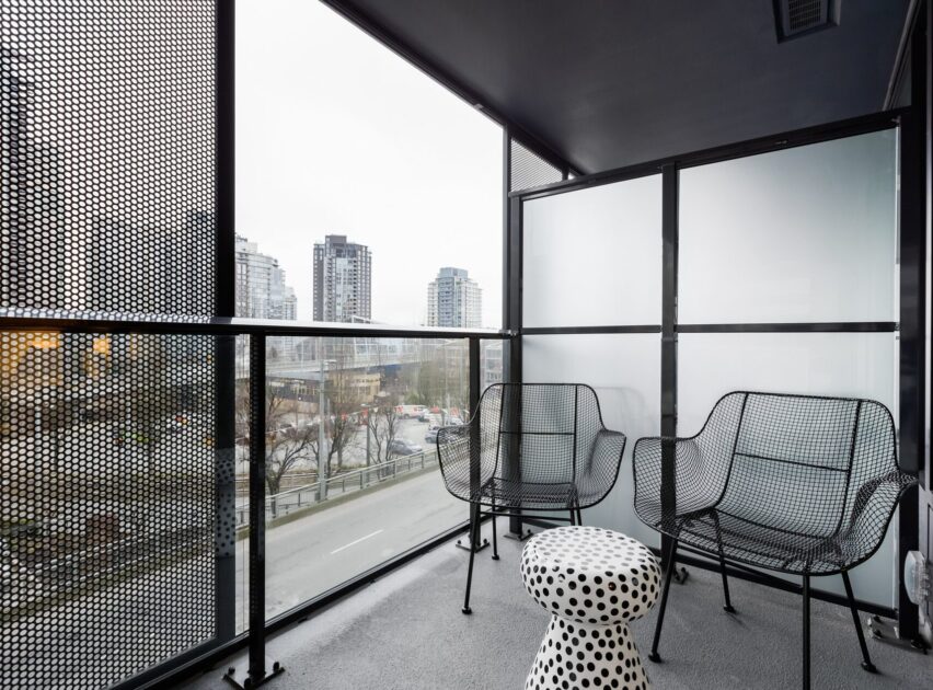 A modern balcony with black mesh chairs, a round polka-dot table, glass and perforated metal railings, and a view of city buildings and a street below.