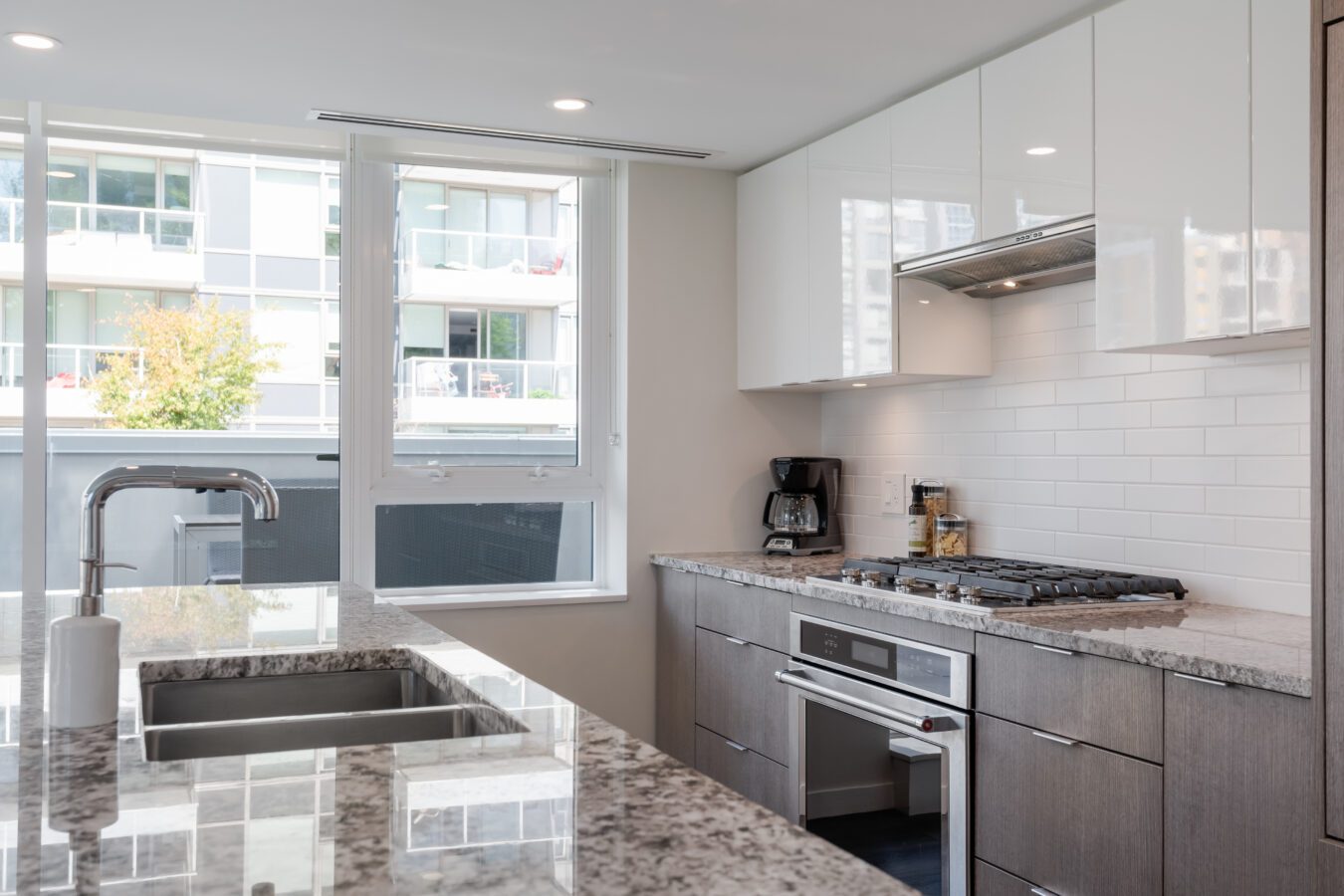 Modern kitchen with granite countertops, a double sink, stainless steel oven and stove, coffee maker, and white cabinets. Large windows provide natural light and a view of an apartment building outside.