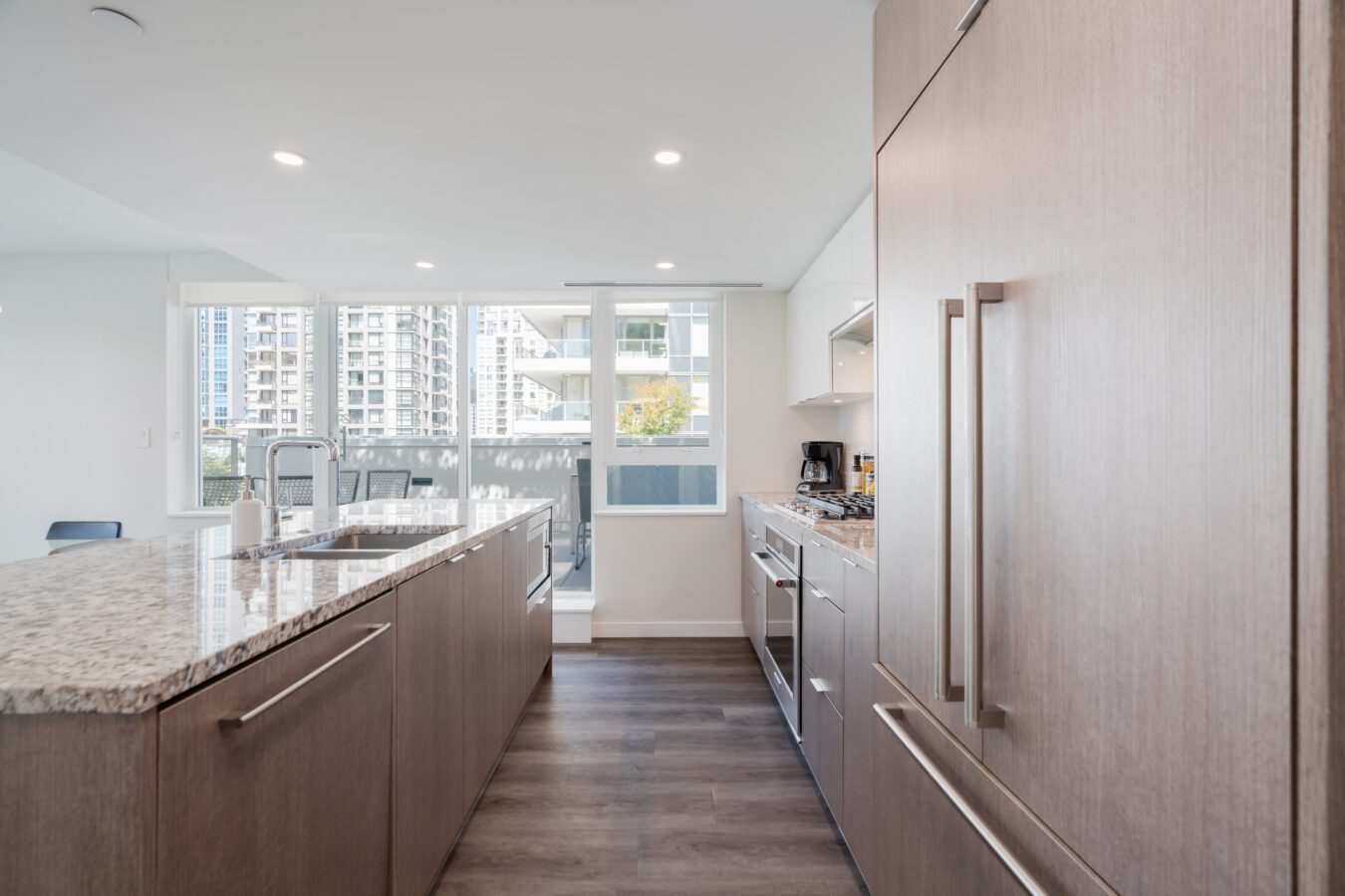Modern kitchen with wood cabinetry, granite island countertop, built-in appliances, and large windows letting in natural light. City buildings are visible outside through the windows.