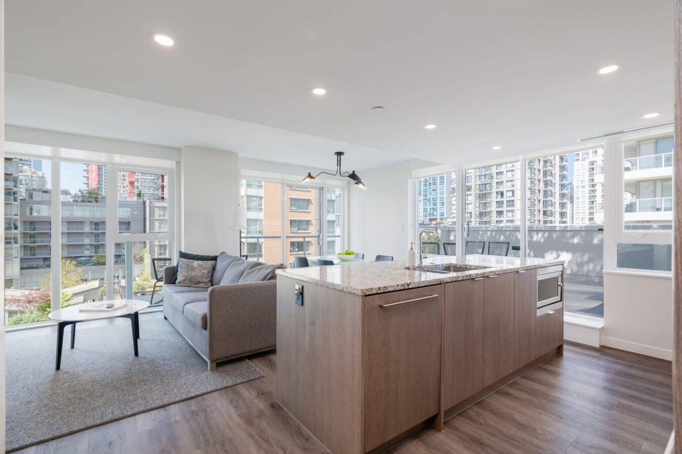 Modern apartment interior with large windows, a gray sofa, coffee table, and open kitchen with a marble island. Urban buildings are visible outside, and natural light fills the space.