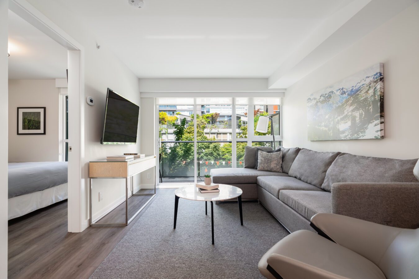 A modern living room with a gray sectional sofa, round coffee table, wall-mounted TV, and large windows letting in natural light. A bedroom with a bed is visible through an open doorway on the left.