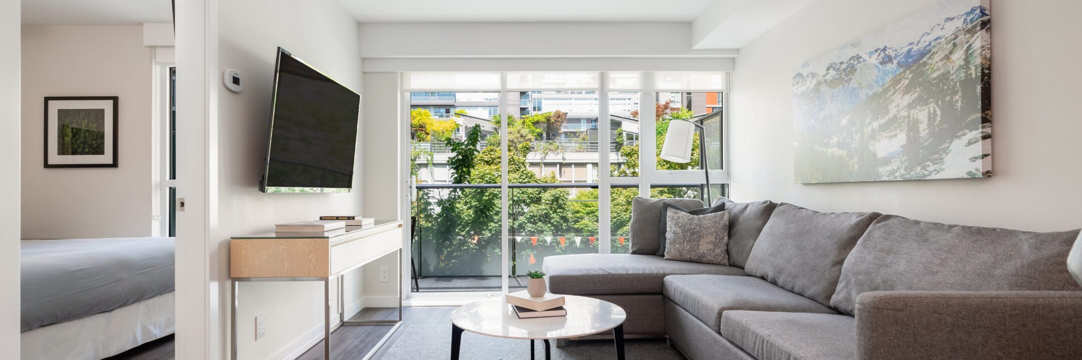 A bright, modern living room with a gray sectional sofa, a round coffee table, and a large wall-mounted TV. Floor-to-ceiling windows let in natural light and offer a view of greenery outside. A bedroom is visible through a doorway.