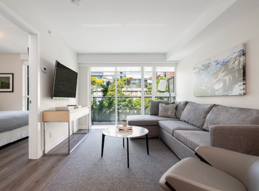 A modern living room with a gray sectional sofa, round coffee table, wall-mounted TV, artwork, and large floor-to-ceiling windows letting in natural light. A glimpse of a bedroom is visible through an open door.
