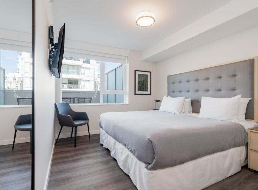 A modern, bright bedroom with a large bed, gray headboard, white and gray bedding, two pillows, two bedside tables, a wall-mounted TV, two chairs by the window, and light wood floors. Large windows let in natural light.