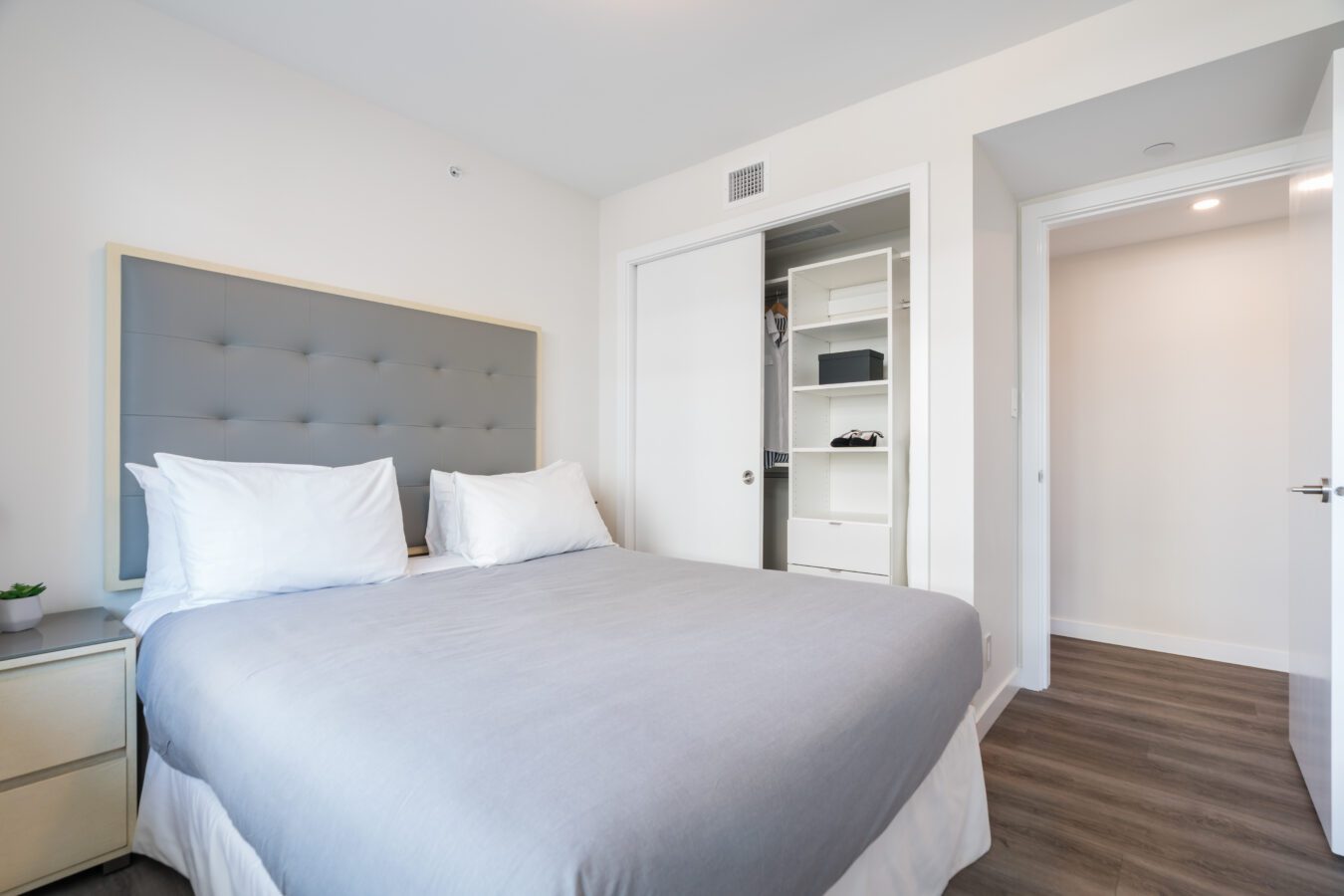 A modern bedroom with a gray upholstered headboard, white bedding, a nightstand with a small plant, an open closet with shelves, and wood flooring. The room has white walls and an open door to a hallway.
