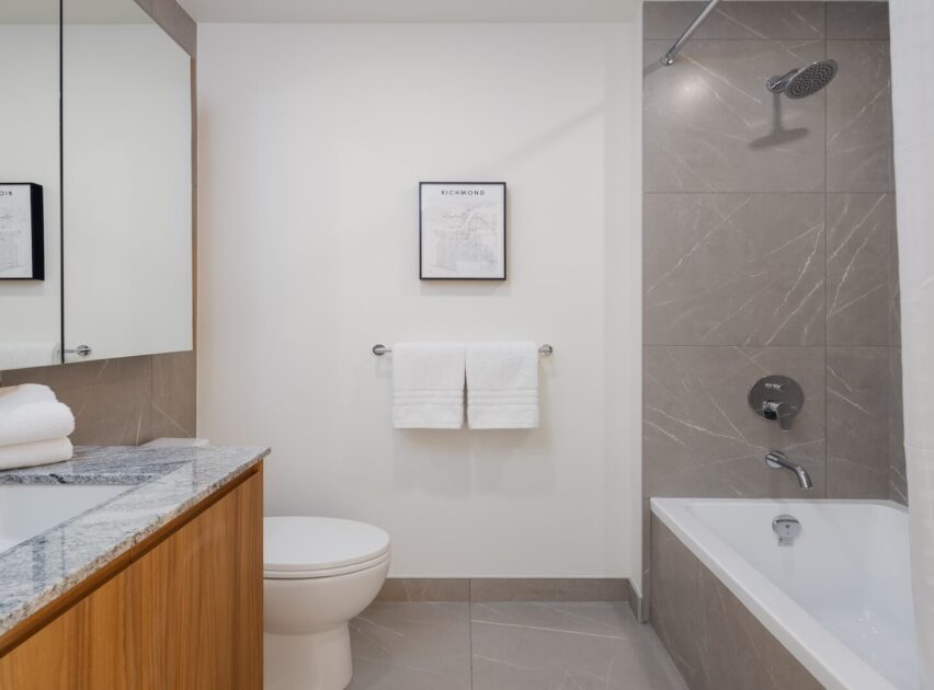 Modern bathroom with a bathtub-shower combo, gray tiles, white walls, a toilet, a vanity with a granite countertop, a wall-mounted towel rack with two white towels, and a framed picture above the towels.