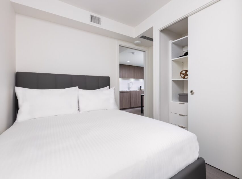 A modern, minimal bedroom with a neatly made bed, white bedding, and a dark headboard. A sliding closet door is open, revealing shelves. The room opens into a kitchen area with wooden cabinets.