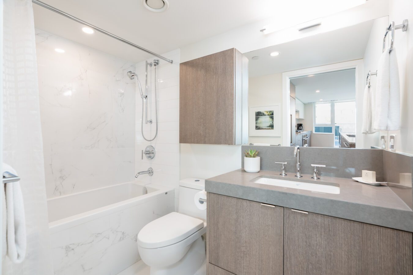 Modern bathroom with a bathtub-shower combo, gray countertop with sink, wooden cabinets, toilet, large mirror, and white walls with marble accents. Towel racks and a small plant add simple decor.