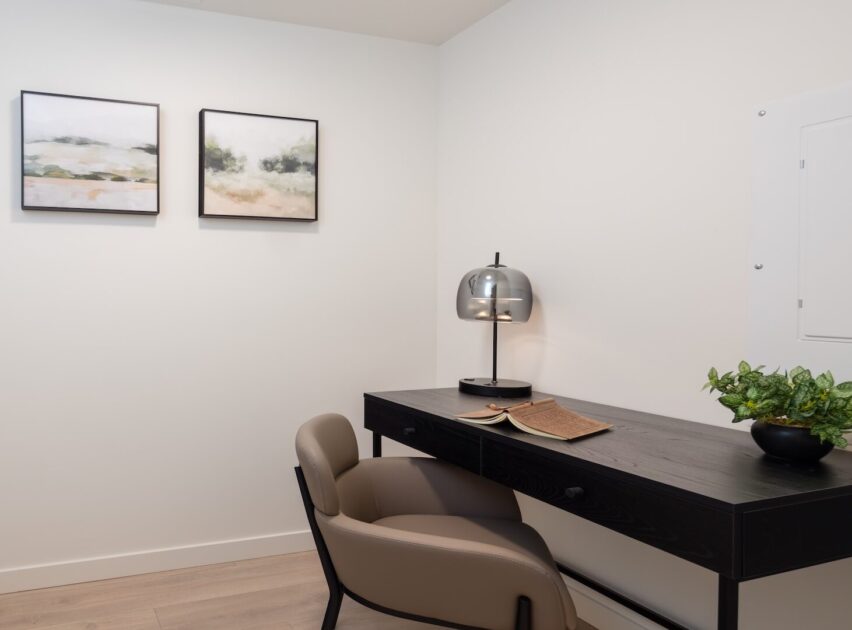 A modern minimalist workspace with a black desk, beige chair, small black lamp, open book, potted plant, and two abstract landscape paintings on a white wall.