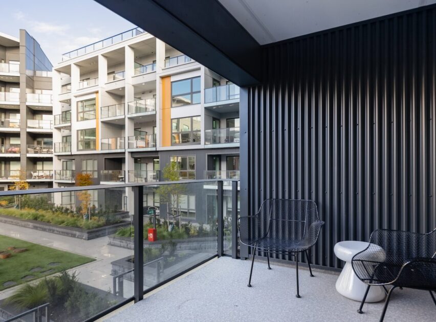 A modern apartment balcony with glass railings, two black chairs, and a small white table overlooks a landscaped courtyard and neighboring contemporary apartment buildings.