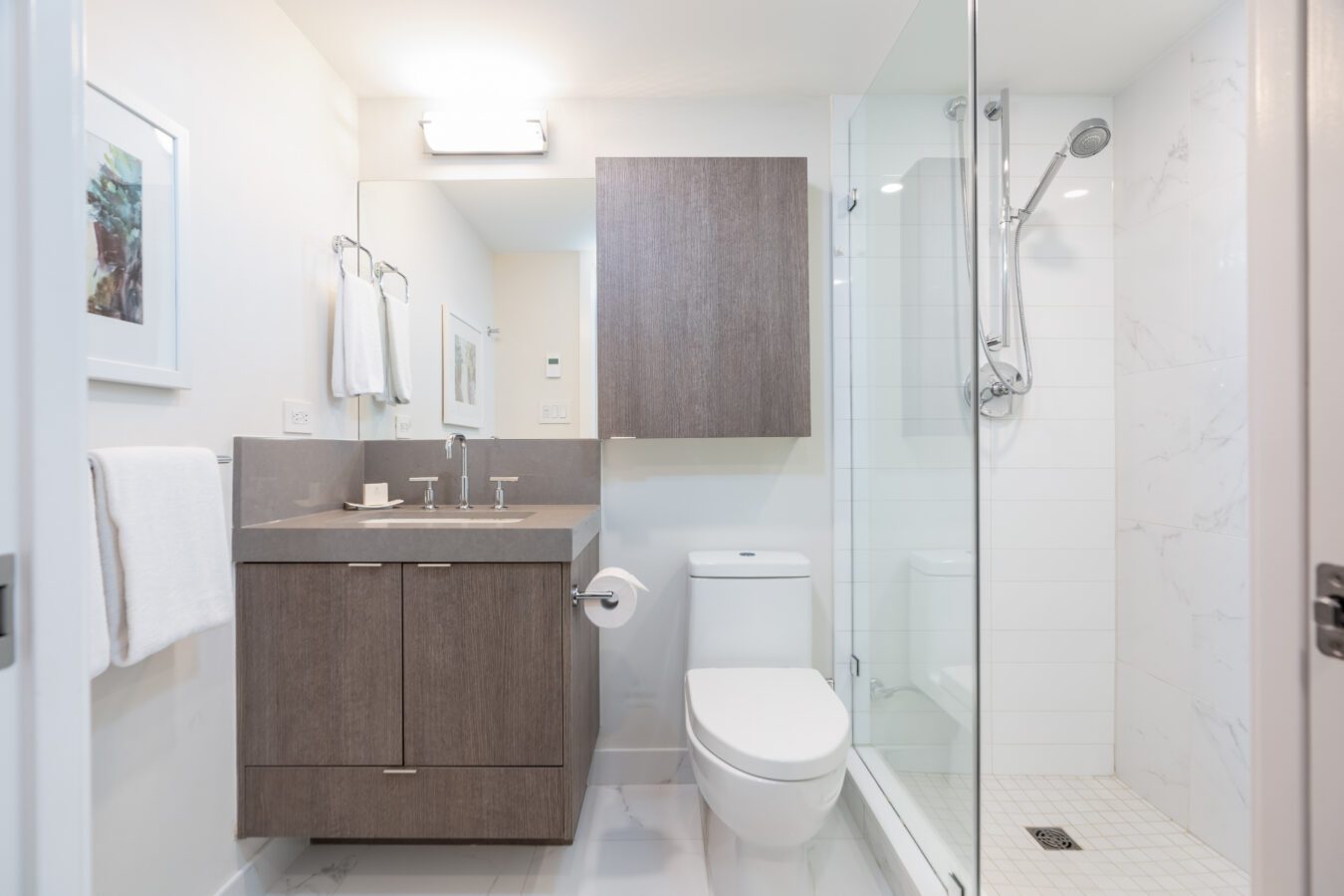 Modern bathroom with a wooden vanity, large mirror, white toilet, glass-enclosed shower, wall-mounted cabinet, towel racks, and light-colored walls and floor. A framed picture hangs on the left wall.