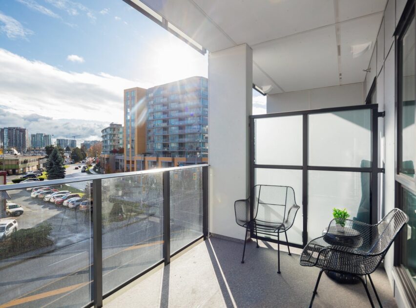 A modern apartment balcony with two black chairs, a small table with a plant, glass railings, and a city view under a bright, sunny sky. An apartment building and parking lot are visible in the background.