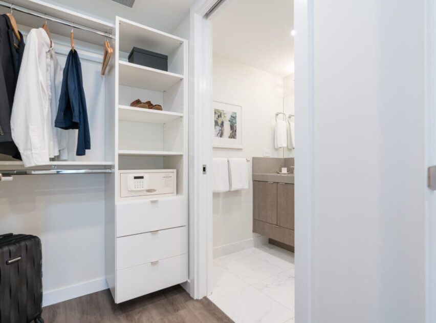 A modern closet with hanging clothes, shelves holding shoes and a safe, and drawers below. To the right, an open door reveals a bright bathroom with towels and a vanity.
