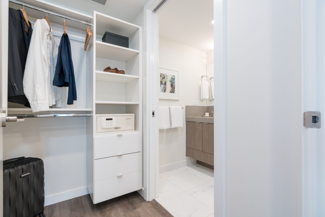A modern closet with hanging clothes, shelves, a safe, and drawers, next to an open door leading to a bathroom with white towels, a large mirror, and light-colored cabinets.