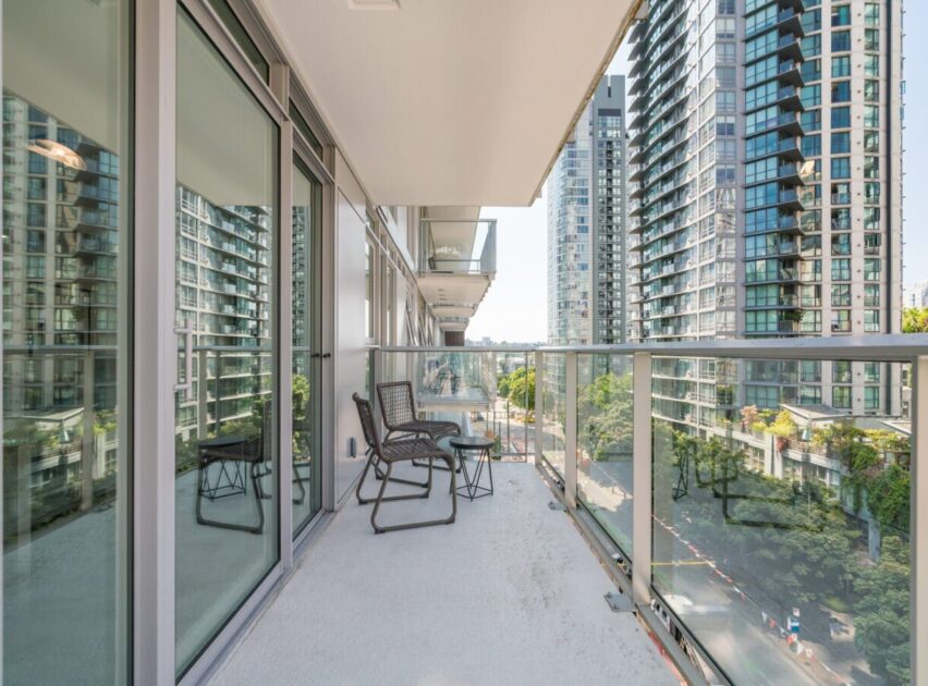 A spacious apartment balcony with glass railings, two chairs, and a small table overlooks a city street lined with modern high-rise buildings and trees on a sunny day.