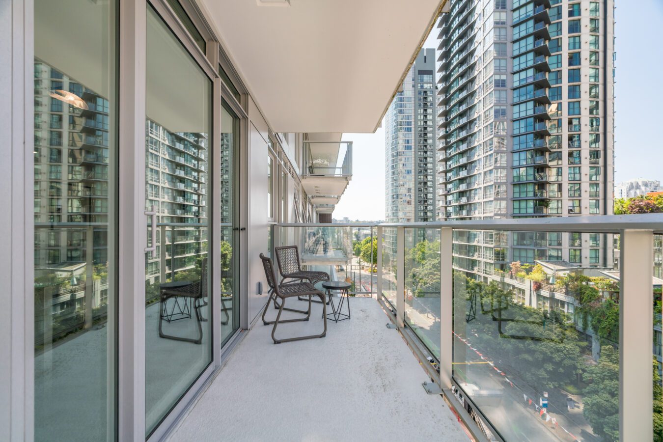 A modern balcony with glass railings features two chairs and a small table, overlooking a street lined with tall glass skyscrapers and lush green trees.